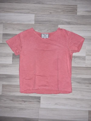 Korallfärgad t-shirt från Denim Base - En enkel och snygg Korallfärgad t-shirt från Denim Base. T-shirten har korta ärmar, rund halsringning och är gjord i mjuk bomull. Perfekt för en avslappnad och trendig stil. Den har ett hål vid lappen i nacken men inget som har stört oss.