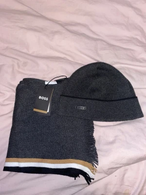 Mörkgrå halsduk och mössa från BOSS - Snygg mörkgrå halsduk sopm har en stilren design med tunna ränder i vitt, svart och beige i ena änden. Halsduken är stickad och har fransar på kortsidorna. Perfekt accessoar.