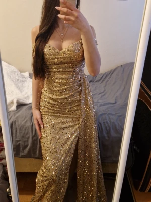 Glittrig guldig långklänning - Säljer en glittrig långklänning i guld med paljetter. Klänningen har offshoulder-modell, markerad midja och en snygg slits framtill. Perfekt för dig som vill sticka ut med en elegant och festlig look.