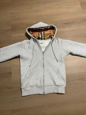 Grå zip hoodie från Burberry - Snygg grå zip hoodie från Burberry med klassiskt rutigt foder i huvan. Hoodien har dragsko i huvan, två fickor fram och ribbade muddar vid ärmslut och nederkant. Perfekt för en chill och stilren look.