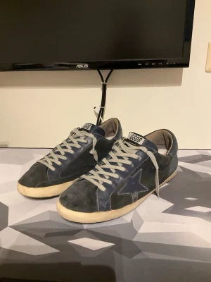 Golden Goose Superstar skor - Snygga mörkblå sneakers från Golden Goose med vit sula och vita skosnören. Skorna har en ikonisk stjärndetalj på sidan och är tillverkade i mocka. GGDB-logga på plösen ger en cool och trendig look. Storlek 42 i fint använt skick.