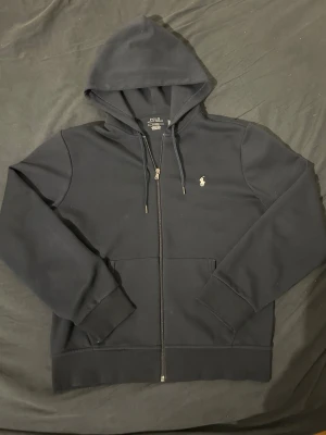 Blå zip tröja från Ralph Lauren - Mörk blå zip tröja från Ralph Lauren med dragkedja framtill och den ikoniska broderade loggan på bröstet. Använd ett fåtal gånger. QR kod finns i tröjan. Nypris: ca 2400