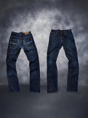 Blå jeans från Nudie Jeans Co W31 L34 - Säljer ett par klassiska blå jeans från Nudie Jeans Co i storlek W31 L34. Jeansen har raka ben, fem fickor och kontrastsömmar i orange. Tillverkade i slitstark bomull med snygg tvätt och detaljer i kopparfärgade nitar.