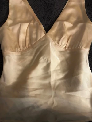 Champagnefärgad satinblus med v-ringning - Snygg champagnefärgad blus i satin med djup v-ringning och breda axelband. Blusen har en figurnära passform och lätt rynkade detaljer vid bysten. Perfekt att styla med jeans eller kjol för en trendig look.