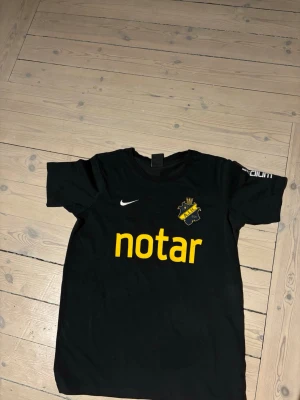 Svart AIK matchtröja från Nike - Svart AIK matchtröja med klubbmärke på bröstet, Nike-logga och gul sponsorlogga 'notar' framtill. Rund halsringning och korta ärmar. Perfekt för fotbollsträning eller att bära på läktaren. Står XL men passar XS eftersom det är barn storlek 