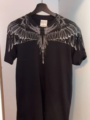 Svart t-shirt Marcelo Burlon med vingar - Svart t-shirt från Marcelo Burlon med ikoniskt vingmönster i vitt och grått över axlar och bröst. Klassisk passform med rund hals och korta ärmar. Perfekt statement-plagg för dig som gillar streetwear.