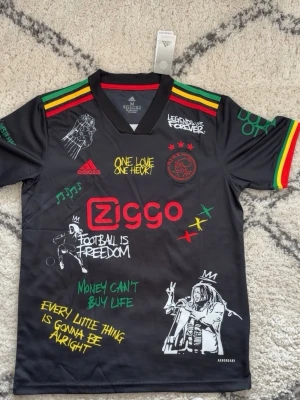 Ajax x Bob Marley svart matchtröja - Svart Ajax matchtröja från Adidas med Bob Marley-tema, färgglada detaljer i rött, gult och grönt på ärmarna, tryckta citat och illustrationer samt Ziggo-logga. Materialet är lätt och andas, perfekt för fotboll eller streetwear.
