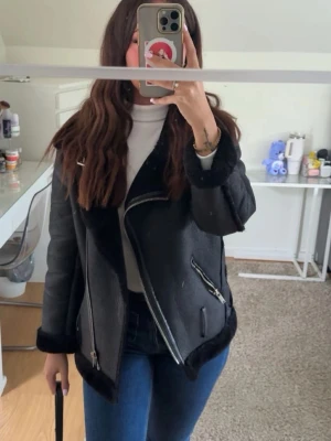 Svart skinnjacka med fuskpälsdetaljer - Snygg svart skinnjacka med fuskpäls på krage och ärmslut. Jackan har sneddragna silverfärgade dragkedjor och en cool bikerstil. Perfekt för dig som vill ha en trendig och stilren look.