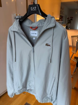 Ljusgrå vindjacka från Lacoste - Snygg ljusgrå vindjacka från Lacoste med dragkedja, huva och två sidofickor. Jackan har det klassiska krokodilmärket på bröstet och meshfoder på insidan för extra komfort. 