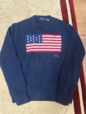 Marinblå stickad tröja Polo Ralph Lauren - Snygg marinblå stickad tröja från Polo Ralph Lauren med amerikansk flagga och RL-broderi på bröstet. Tröjan har rund halsringning, ribbstickade muddar och är gjord i mjukt bomullsmaterial. Perfekt för dig som gillar klassisk preppy stil.