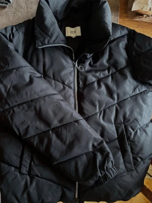 Svart pufferjacka från JDY - Snygg svart pufferjacka från JDY med dragkedja framtill och två sidofickor. Jackan har quiltat mönster, hög krage och elastiska muddar vid ärmsluten. Perfekt för kalla vinterdagar och ger en stilren look.