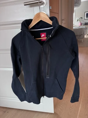 Svart Nike hoodie med dragkedja - Svart hoodie från Nike med hel dragkedja framtill och en extra bröstficka med dragkedja. Klassisk huva med dragsko och två sidofickor. Tillverkad i mjukt sweatshirtmaterial, perfekt för en avslappnad och sportig stil. Junior S, Vuxen XS