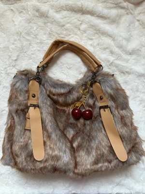 Beige fluffig handväska med körsbär - Säljer en beige och brun fluffig handväska med fejkpäls och ljusbruna detaljer i konstläder. Väskan har dubbla handtag och en söt körsbärsnyckelring som dekoration. Insidan är fodrad i brunt tyg och har en innerficka för småsaker.