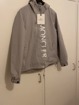 Grå vindjacka från M****er - Snygg grå vindjacka från Moncler med stor vit logga längs dragkedjan. Jackan har huva, tre fickor med dragkedja och justerbar nederkant. Perfekt för blåsiga dagar och stilrena outfits. Materialet är lätt och vattenavvisande.