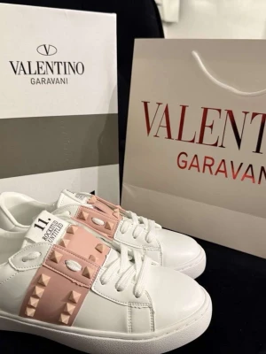 Valentino Garavani sneakers med nitar - Snygga vita sneakers från Valentino Garavani med breda ljusrosa band över vristen dekorerade med ikoniska pyramidformade nitar. Skorna har klassisk låg profil, rund tå och vita snören. Tillverkade i slätt läder för en clean och lyxig look.