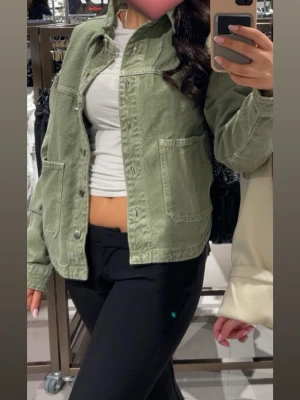 Olivgrön jeansjacka med fickor - Snygg olivgrön jeansjacka med två stora framfickor och klassisk krage. Jackan har knappstängning framtill och avslappnad passform, perfekt att slänga över en t-shirt. Passar dig som gillar en enkel men trendig look. Jackan är snålt använd och har inga skador. 