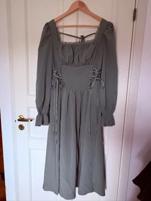 Grön långklänning med snörning - Snygg grön långklänning med puffiga långärmar och markerad midja med snörningsdetaljer på sidorna. Fyrkantig ringning och volang vid ärmslut ger en romantisk vibe. Ser mer grå ut på bild.