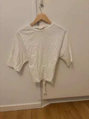 Vit croppad topp från Nelly - Snygg vit croppad topp från Nelly med knyt i midjan. T-shirten har korta ärmar och rund halsringning. Perfekt att matcha med jeans eller kjol för en fräsch look. Använd en gång, den är nyskick 