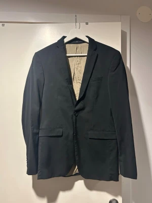 Svart kostym med kavaj och byxor - Säljer en klassisk svart kostym med enkelknäppt kavaj och matchande byxor. Kavajen har två framfickor med lock, bröstficka och knappar vid ärmslut. Fodrad insida i beige. Perfekt för dig som vill ha en stilren och tidlös look.