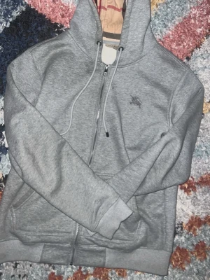 Grå hoodie med dragkedja från Burberry - Snygg grå hoodie från Burberry med dragkedja framtill och justerbar huva. Broderad logga med enhörning på bröstet. Mjuk bomullsblandning och ribbade muddar vid ärmslut och nederkant. Perfekt för en chill och stilren look.