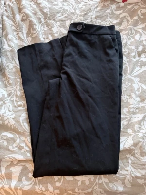 Svarta kostymbyxor med knapp/ slits i benen  - Snygga svarta kostymbyxor med rak passform och klassisk knappstängning i midjan. Byxorna har diskreta fickor och är perfekta för en clean och stilren look. En mjuk och följsam textil som ger en dressad känsla.