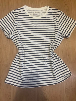 Randig t-shirt från PSBN Sister - Säljer en klassisk vit t-shirt med mörkblå horisontella ränder från PSBN Sister. T-shirten har rund halsringning och korta ärmar. Tillverkad i mjuk bomull, perfekt för en avslappnad och stilren look.