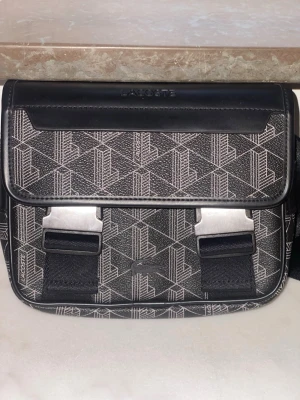 Lacoste väska - Snygg svart axelväska från Lacoste med geometriskt mönster i vitt och detaljer i svart konstläder. Väskan har justerbar axelrem och två silverfärgade spännen framtill. Perfekt för dig som vill ha en stilren och modern accessoar, väskan köptes från zalando för 2000kr och har bara använts 1 gång pris kan diskuteras vid snabb affär 