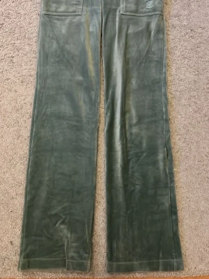 Gröna velourbyxor Juicy Couture - Mysiga gröna mjukisbyxor i velour från Juicy Couture med raka ben, bred resår i midjan och dragsko. Byxorna har två stora fickor framtill och en broderad JC-logga på ena fickan. Perfekta för en chill och trendig look.