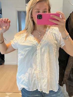 Vit spetsblus med korta ärmar - Säljer en vit blus med korta ärmar och vacker spetsdetalj längs knäppningen och halsringningen. Blusen har en luftig passform och är gjord i ett tunt, lätt material som känns fräscht och somrigt. Perfekt att matcha med jeans eller kjol.