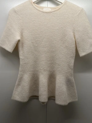 Krämvit stickad topp med volang - Supersöt krämvit stickad topp med korta ärmar och volang nedtill. Ribbstickad struktur och rund halsringning ger en clean look. Perfekt att styla med jeans eller kjol för en sopistikerad vibe. Materialet känns mjukt och mysigt. Har bara testat varan en gång