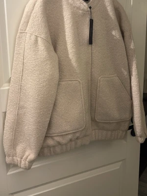 Beige teddyjacka med stora fickor - Mysig beige teddyjacka med dragkedja framtill och två stora fyrkantiga fickor. Jackan har resår vid ärmslut och nederkant för en avslappnad passform. Tillverkad i fluffigt teddy-material som ger en trendig och varm look.