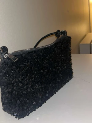 Svart paljettkuvertväska - Snygg svart kuvertväska täckt med glittrande paljetter och ett kort handtag. Väskan har en rektangulär form och stängs med dragkedja i silverfärg. Perfekt accessoar för att lyfta din outfit med! Materialet är tyg täckt av paljetter.