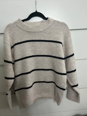 Beige stickad tröja med svarta ränder - Snygg beige stickad tröja med svarta horisontella ränder. Tröjan har en bred ribbad rundhalsad krage och långa ärmar med ribbade muddar. Perfekt för lager på lager och enkel att matcha till olika outfits. TRÖJAN ÄR STOR I STORLEKEN!!