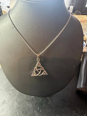 Silvrigt halsband med keltisk triangel - Snyggt halsband i silverfärgad metall med en keltisk triangelberlock. Berlocken har ett flätat mönster och hänger på en kedja. Perfekt för dig som gillar unika och mystiska smycken med symbolik.