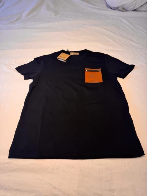 Svart Burberry t-shirt med läderdetalj - Snygg svart t-shirt från Burberry med en unik brun läderficka på bröstet, dekorerad med Burberrys klassiska ryttare och texten 'London'. T-shirten har rund hals och korta ärmar. Perfekt för dig som gillar exklusiva detaljer och stilren design.