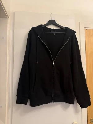 Svart hoodie med dragkedja från H&M - Klassisk svart hoodie från H&M med dragkedja framtill, två fickor och justerbar huva med snören. Regular fit-modell i mjukt sweatshirtmaterial som passar perfekt till jeans eller joggers. Enkel och stilren design utan tryck eller mönster. Inge tecken på användning, inga skador osv.