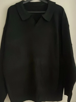 Svart stickad tröja med krage - En stilren svart stickad tröja med klassisk krage och v-ringning. Tröjan har långa ärmar och ribbade muddar vid ärmslut och nederkant. Perfekt för dig som gillar en enkel men snygg look.