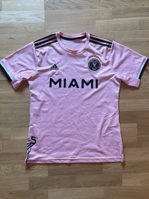 Rosa Inter Miami matchtröja Adidas - Säljer en rosa Inter Miami matchtröja från Adidas med svarta detaljer och klubbmärke på bröstet. Tröjan har korta ärmar, rund hals och svarta ränder på axlarna. Materialet är lätt och andas, perfekt för träning eller match.
