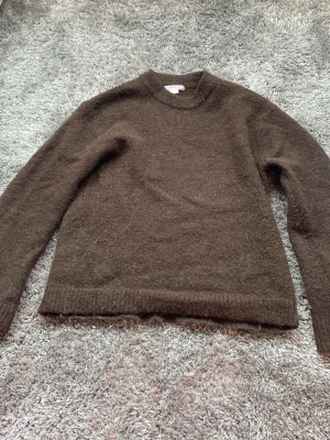 Brun stickad tröja Gina Tricot - Mysig brun stickad tröja från Gina Tricot med rund halsringning och långa ärmar. Tröjan har en fluffig yta och ribbstickade muddar vid ärmslut och nederkant. Perfekt för kyliga dagar och enkel att matcha med olika outfits.