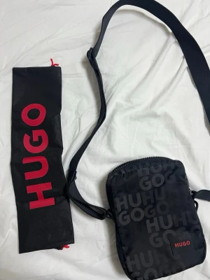 Svart axelväska från HUGO - Snygg svart axelväska från HUGO med justerbar rem och dragkedja. Väskan har ett ton-i-ton HUGO-mönster över hela ytan och en liten röd HUGO-logga framtill. Tillverkad i slitstarkt syntetmaterial och kommer med en svart dustbag med röd logga.