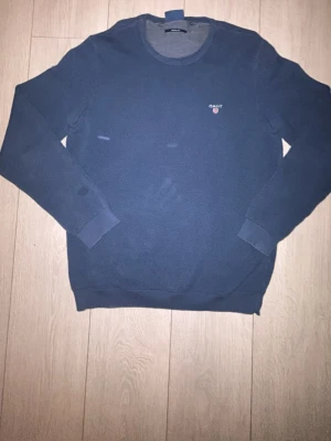 Mörkblå tröja från GANT - Mörkblå långärmad tröja från GANT med rund halsringning och diskret logga broderad på bröstet. Tröjan har ribbade muddar vid ärmslut och nederkant. Perfekt för en stilren och enkel look.