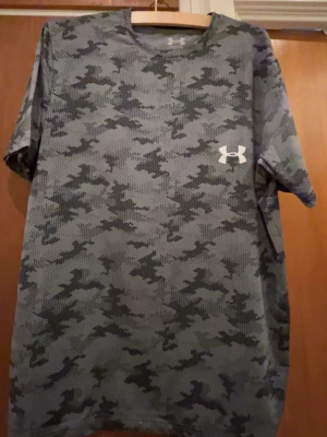Camo t-shirt från Under Armour - Säljer en grå och svart camomönstrad t-shirt från Under Armour. T-shirten har rund hals, korta ärmar och är tillverkad i ett lätt och ventilerande soprtmaterial. Perfekt för dig som gillar sportig stil och streetwear.