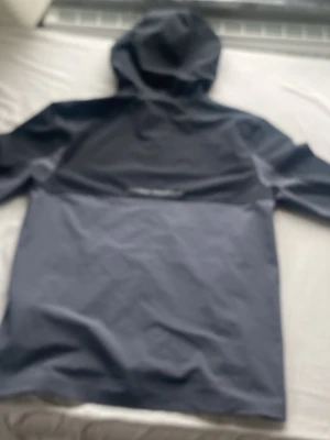 Svart och grå vindjacka från Under Armour - Snygg vindjacka från Under Armour i svart och grått med huva och dragkedja framtill. Jackan har logga på bröstet och är tillverkad i ett lätt, vattenavvisande material som passar perfekt för blåsiga dagar.