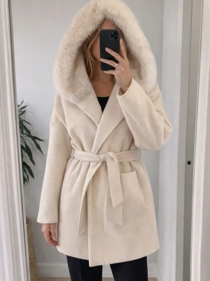 Beige kappa med pälsdetaljer - Elegant beige kappa med stor huva och pälsdetaljer. Kappan har ett knytbälte i midjan och praktiska fickor framtill. Perfekt för kyliga dagar när du vill hålla stilen. 