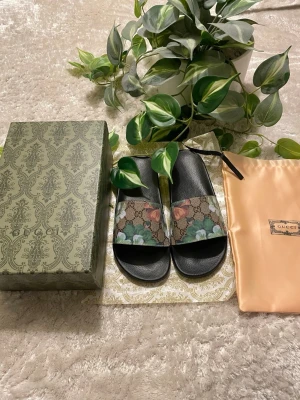 Gucci sandaler med blommönster - Säljer ett par snygga sandaler från Gucci med svart sula och bred rem med klassiskt GG-mönster och blommor i grönt, vitt och rosa. Sandalerna kommer i originalkartong och med dustbag. Perfekt för dig som vill ha något exklusivt och trendigt.