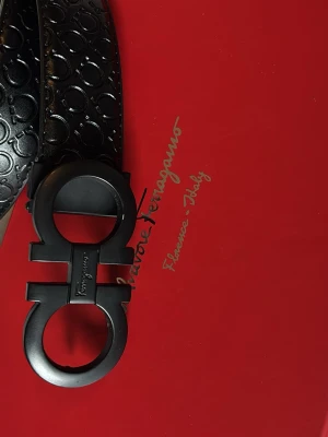 Svart bälte från Salvatore Ferragamo - Snyggt svart bälte från Salvatore Ferragamo med präglat mönster och ikoniskt dubbel-Gancio-spänne i matt svart metall. Bältet har en exklusiv känsla och passar perfekt till både jeans och kostymbyxor. Ett statement-accessoar för dig som gillar lyxiga detaljer.