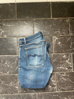 Blå slitna jeans från Nudie Jeans - Säljer ett par blå jeans från Nudie Jeans med slitningar och hål på knäna. Klassisk femficksmodell med ljus tvätt och smal passform. Snygga detaljer på bakfickan och contast-sömmar. Perfekta för en avslappnad streetstil.