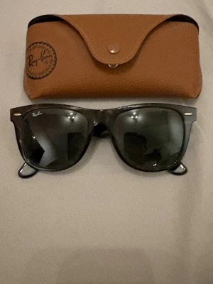 Ray-Ban svarta solglasögon - Säljer ett par klassiska svarta solglasögon från Ray-Ban med ikonisk logga på bågen. Glasögonen har en tidlös form och kommer med ett snyggt brunt fodral i skinn. Perfekt för dig som vill ha en stilren accessoar. Sitter bra på ny pris ca 1500kr. Mitt pris kan diskuteras vid snabb affär.