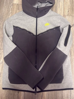 Nike Tech Fleece hoodie grå/svart - Nike Tech Fleece hoodie i grått och svart med hel dragkedja och huva. Snygg kontrastdesign med svart V-formad panel framtill och svart detalj på ärmarna. Liten neon Nike-logga på bröstet och ficka med dragkedja på ena ärmen. Perfekt för en sportig och modern look.
