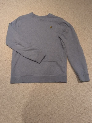 Blå sweatshirt från Lyle & Scott - Säljer en stilren blå sweatshirt från Lyle & Scott med deras klassiska gul fågel-logga på bröstet. Tröjan har rund hals, långa ärmar och ribbade muddar. Perfekt för en avslappnad look. Helt oanvänd/Ny.